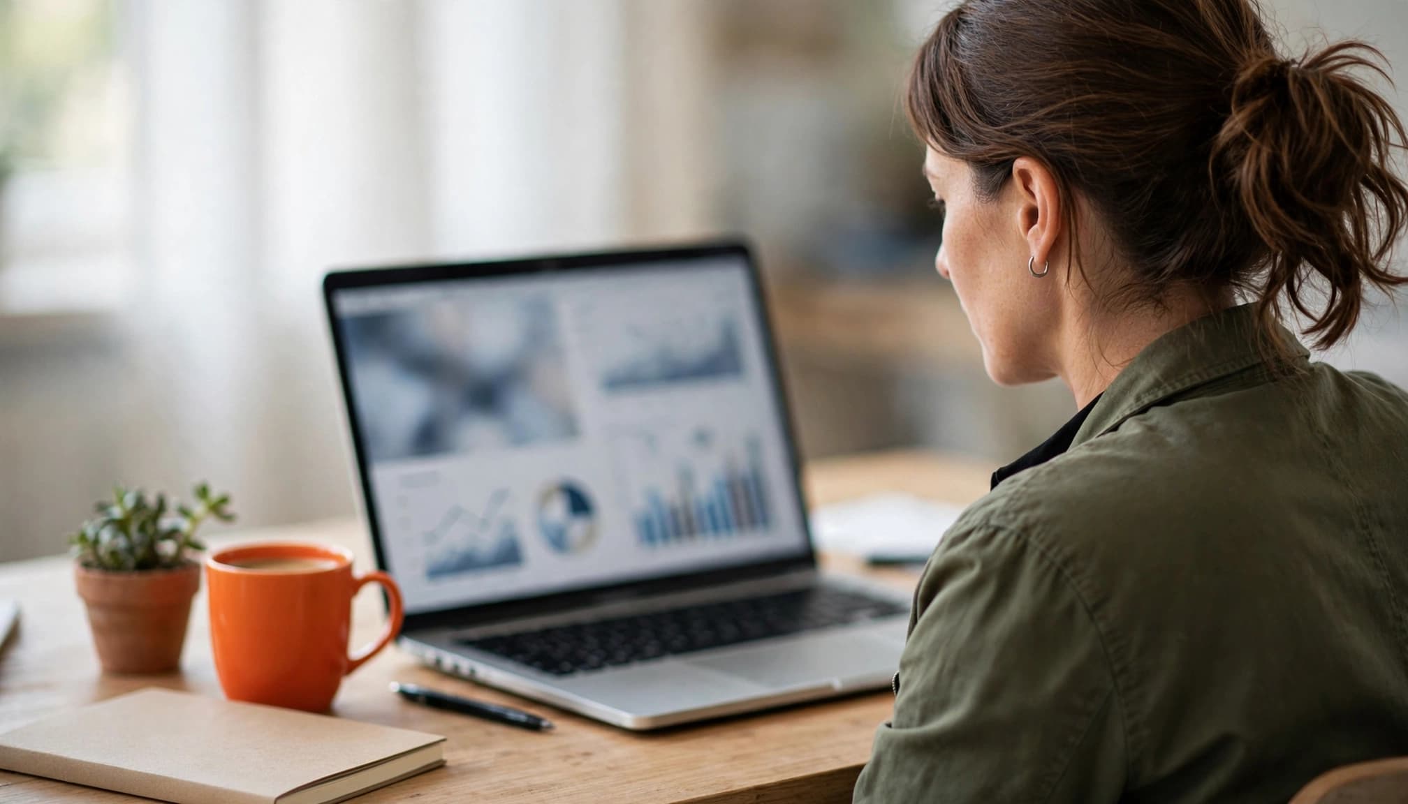 A business owner reviews blurred charts on a laptop beside coffee, a notebook, and a plant.